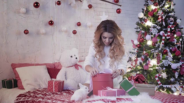 年轻漂亮的女人坐在圣诞树旁的床上，包裹着一份新年礼物视频素材
