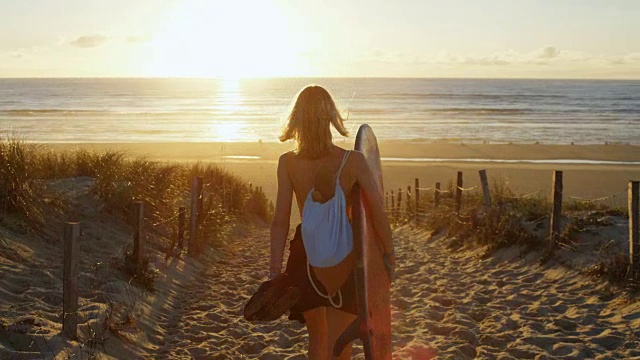 美丽的年轻女子在一个泳衣拿着冲浪板在海滩上走向大海。阳光照耀着这个美丽的女孩。海是可见的。视频素材