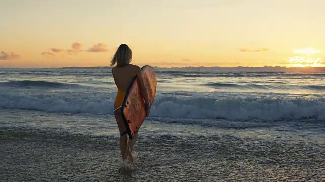 穿着泳衣的美丽年轻女子背着冲浪板走进大海。她看起来美丽和苗条，有一头金发。大海浪和日落的背景。视频素材