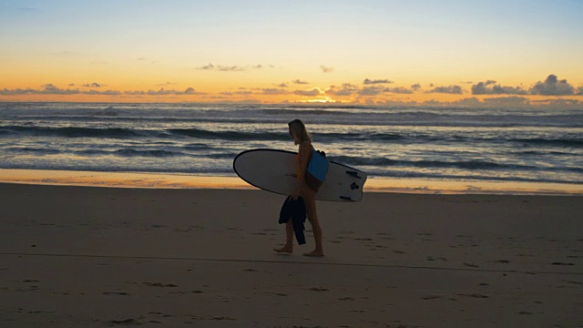 侧视图镜头美丽的年轻女子在泳装沿着海滩行走，而携带冲浪板。海与波浪和日落的背景。缓慢的运动。视频素材