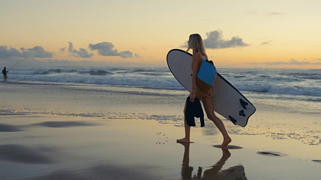 穿着泳衣的漂亮年轻女子背着冲浪板沿着海滩散步。海与波浪和日落的背景。缓慢的运动。视频素材