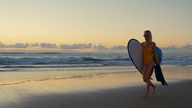 穿着泳衣的漂亮年轻女子背着冲浪板沿着海滩散步。海与波浪和日落的背景。慢动作拍摄。视频素材