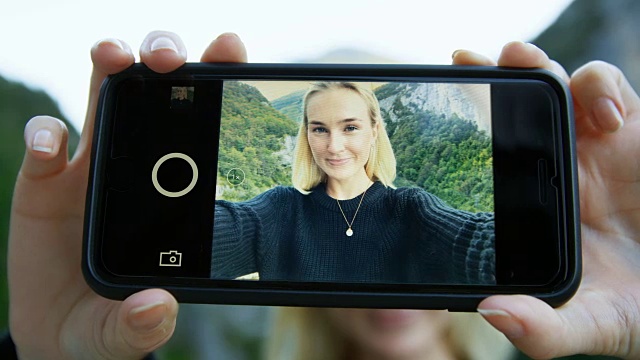 美丽的金发女孩自拍的智能手机屏幕上的特写。在背景风景优美的青山和山景。视频下载
