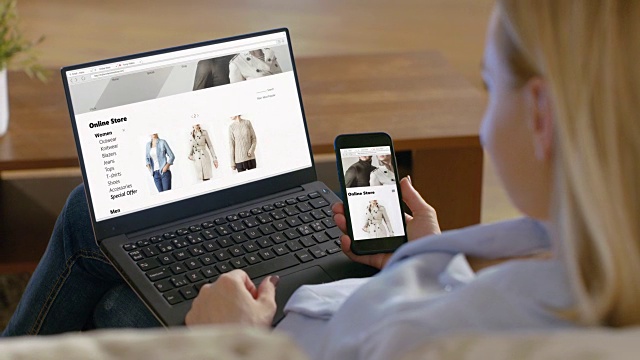 一个女人拿着智能手机，膝盖上放着笔记本电脑，在网上浏览销售时尚服装的商店。视频素材