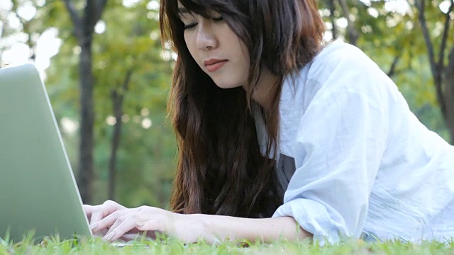 年轻的亚洲女人的腿在绿色草地上与打开的笔记本电脑。女孩的手放在键盘上。远程学习的概念。快乐的年轻亚洲潮女在公园里用笔记本电脑工作。学习在户外。视频下载