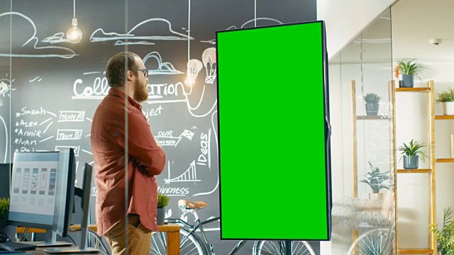 男性开发者看着带有绿色屏幕模型的交互式白板。他捋着胡子思考解决问题的方法。在背景创意办公室与黑板墙与项目计划绘制。视频素材