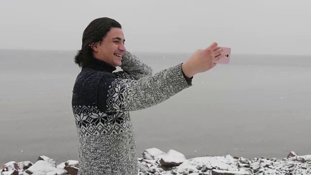 混血的拉丁裔游客在毛衣上自拍在雪海背景。一名男子用智能手机自拍。冬天的雪正在前景下视频素材