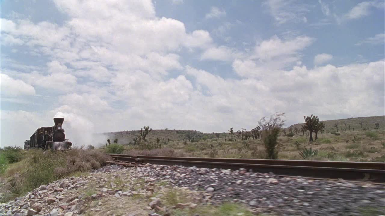 在沙漠或干旱地区，火车沿着铁轨行驶的中等角度。看见白色的蒸汽从火车的发动机里冒出来。周围有绿树、灌木和仙人掌。在背景中看到小山丘或山脉。视频下载