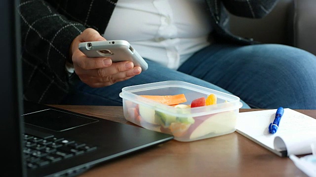 正在节食的商务女性穿着套装，吃着午餐盒里的健康水果零食，看着智能手机视频素材