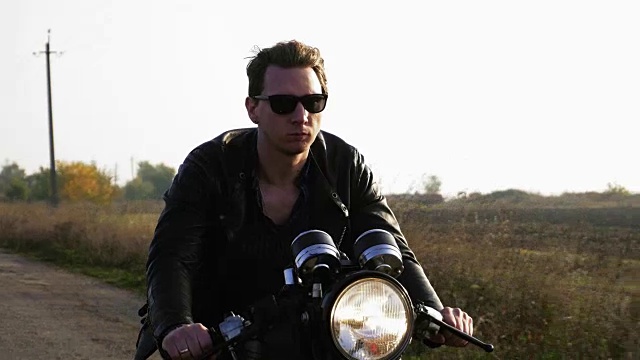 一个时尚酷的年轻人在一个阳光明媚的日子里戴着墨镜和皮夹克骑摩托车在柏油路上的特写视频素材