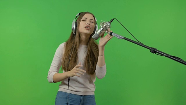 一个小女孩正对着录音室的麦克风唱歌。在绿色的背景上。视频素材