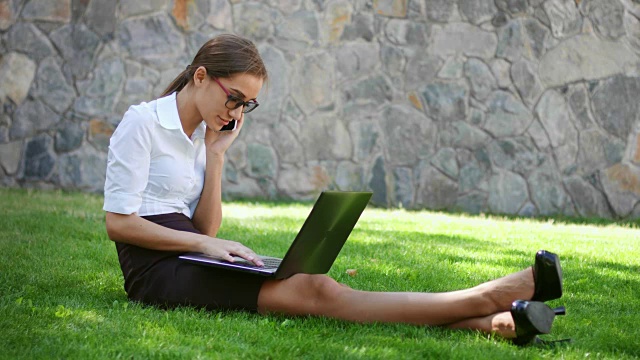 女商人坐在公园的草地上，拿着笔记本电脑，接电话视频素材