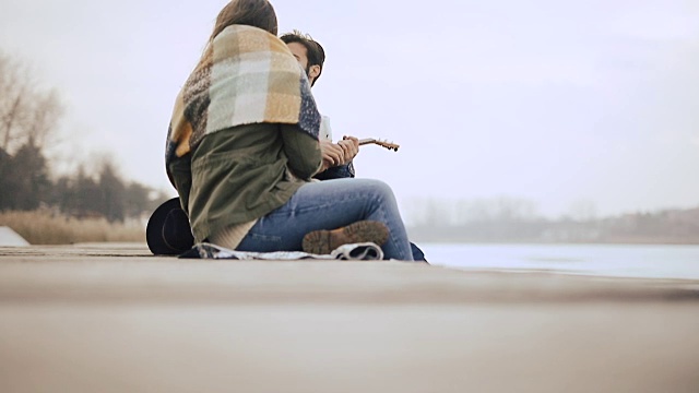 一个年轻人坐在结冰的湖边的码头上给他的女朋友弹奏尤克里里琴视频素材