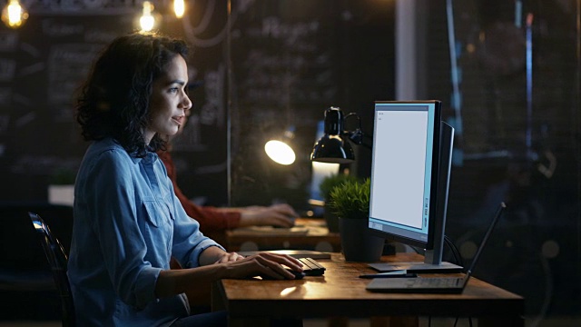 美丽的女性程序员在她的个人电脑上工作。她的男同事坐在她旁边。晚装办公室有创意照明。视频素材