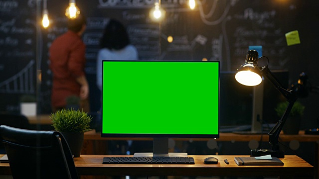 个人电脑与模拟绿色屏幕显示器站在办公桌上，在后台，人们在黑板墙上工作在创意办公室。视频素材