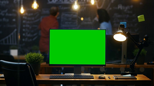 个人电脑与模拟绿色屏幕显示器站在办公桌上，在后台，人们在黑板墙上工作在创意办公室。间隔拍摄。视频素材