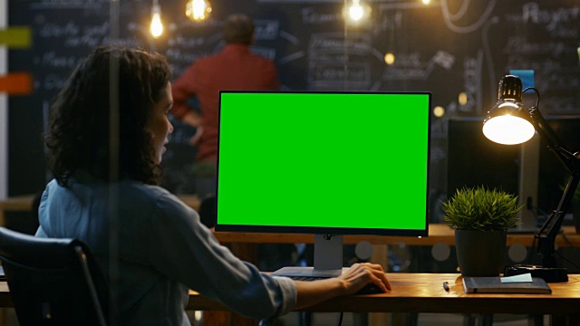 美丽的女办公室职员在她的桌面工作在一个模拟的绿屏个人电脑。过肩镜头。她的同事在后台工作。晚上创意办公室。视频素材