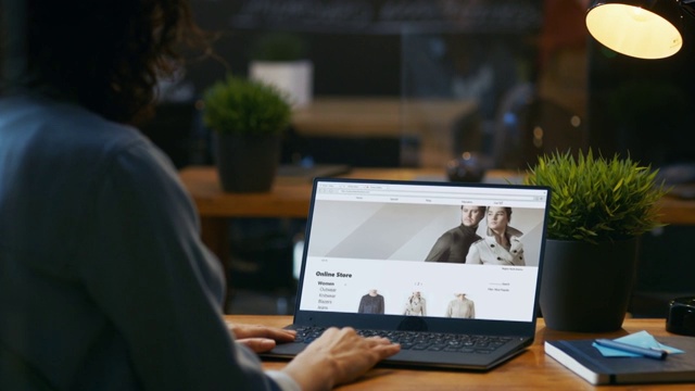一位坐在办公桌前用笔记本电脑浏览服装零售网站的女士。网站展示各种男性和女性时尚的衣服，新系列。视频素材