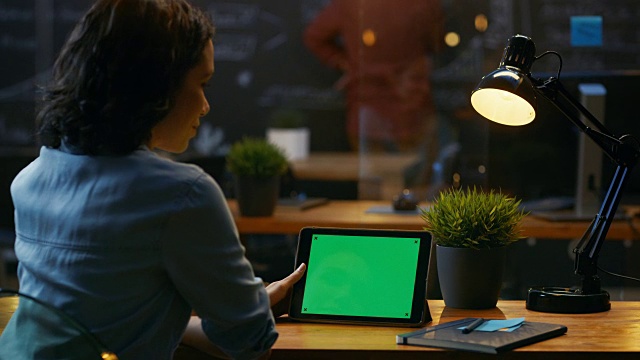 年轻女子坐在她的木制书桌前，使用平板电脑和模拟的绿色屏幕在风景视图。视频素材