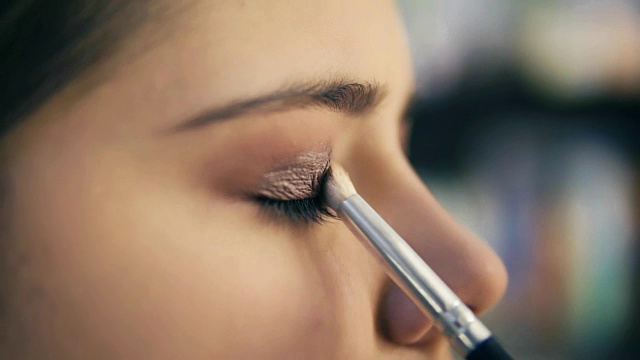 专业化妆师用化妆刷涂抹眼影的手的特写视图。Pro visagiste将浅棕色的眼影涂在模特的眼睑上。Slowmotion拍摄视频购买