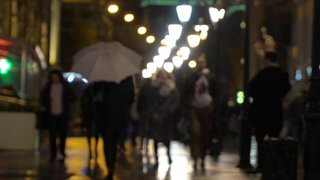 夜晚的城市下雨。雨夜的城市街道上有路灯，沥青路从雨中闪闪发光。不认识的行人，年轻人，拿着白色雨伞的女孩。购物休闲概念，季节，天气，现代城市生活方式视频素材