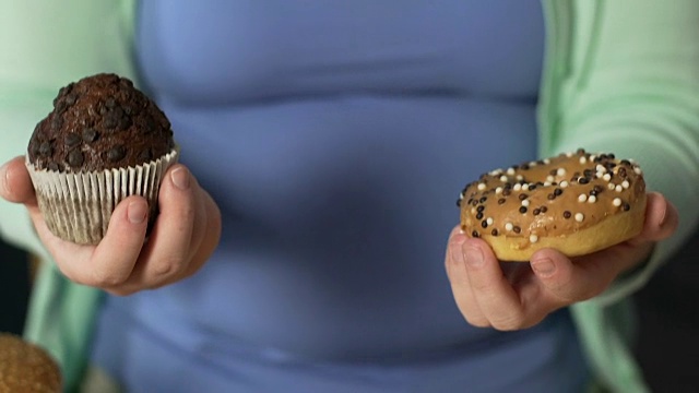 胖乎乎的女性手里拿着松饼和甜甜圈，对甜食上瘾，问题视频素材