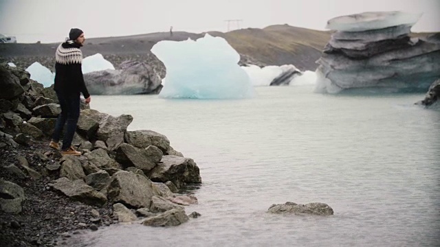 年轻英俊的男人走在冰湖上。快乐的游客坐在岩石上观看冰岛的冰川视频素材