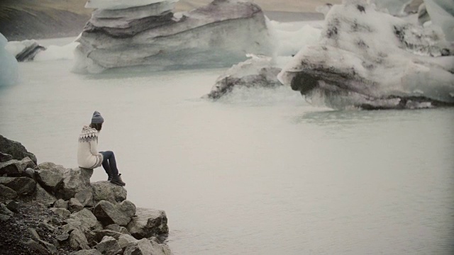 年轻迷人的女人坐在冰泻湖的岩石上。游客在lopapeysa探索冰岛的风景视频素材