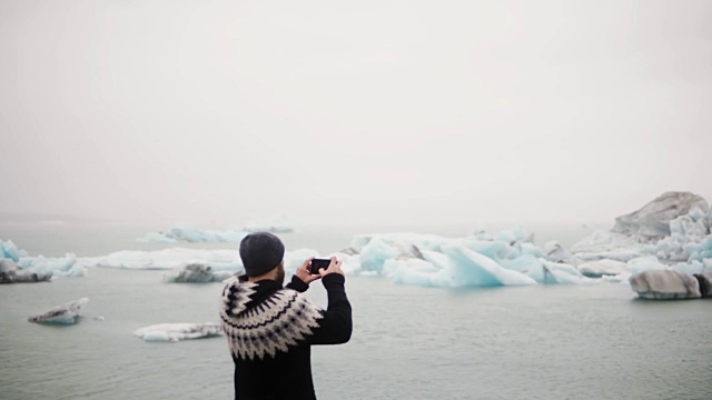 年轻的旅行男子站在冰岛的Jokulsalon冰礁湖，用智能手机拍摄冰川的背影视频素材