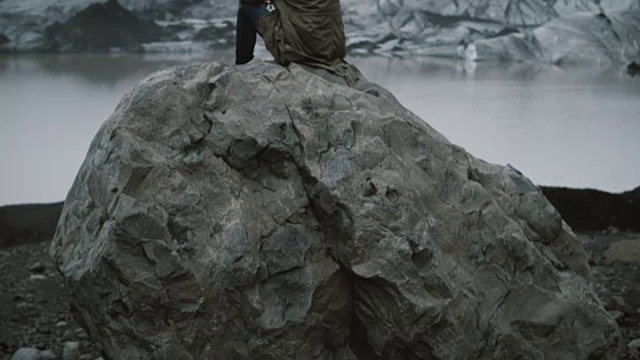 年轻男子独自坐在火山岩上，看着冰岛的冰泻湖冰川的背影视频素材