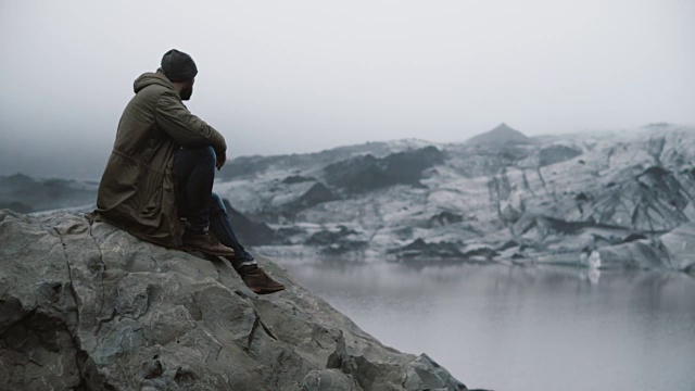 年轻英俊的男子坐在岩石上看着冰川。游客独自探索冰岛的冰泻湖视频素材