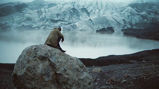 年轻英俊的男子坐在冰泻湖的岩石上看着冰岛的冰川。游客站起来走开视频素材