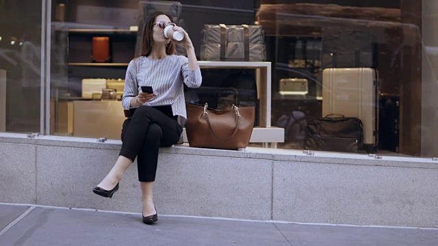 年轻漂亮的女商人坐在橱窗附近，喝着咖啡，在休息时间用着智能手机视频素材