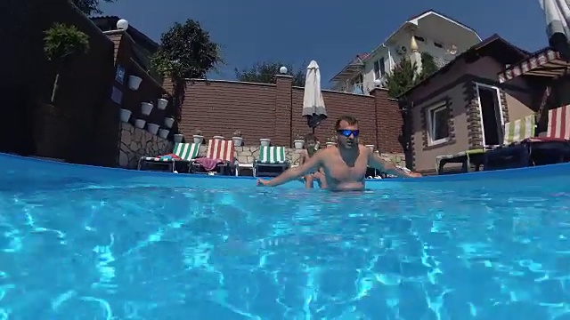 快乐英俊的年轻人在游泳池游泳，水下观，挥挥手。夏天的假期时间。行动的相机视频下载