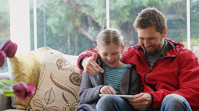 快乐的爸爸和女儿坐在沙发上用他的平板电脑4K 4K视频素材