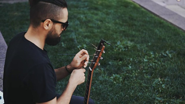 年轻英俊的大胡子男子坐在公园的草地上，在外面换弦，重新弹着吉他视频素材
