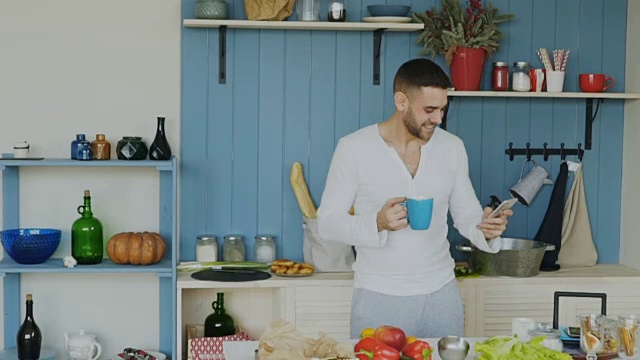 慢动作英俊的年轻有趣的男人跳舞和唱歌在厨房，而在他的智能手机上浏览社交媒体在早上在家里视频素材