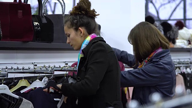 两个年轻漂亮的女孩在一家时尚精品店挑选衣服。女孩们仔细地看衣服，检查它。视频素材