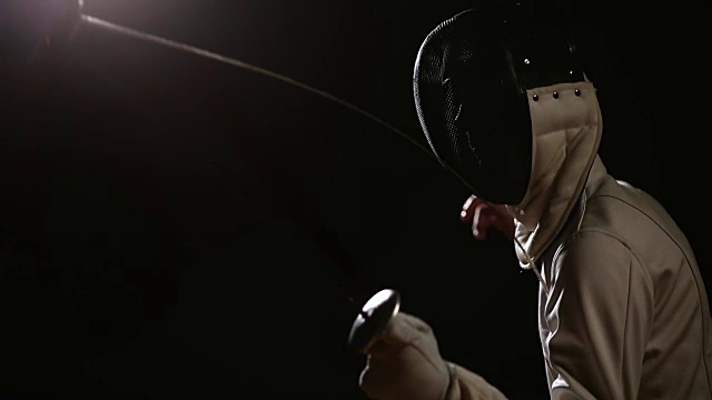 一个男人的特写镜头戴着一个头盔的击剑格斗与一个对手在一个黑暗的背景与剑。工作室拍摄视频下载