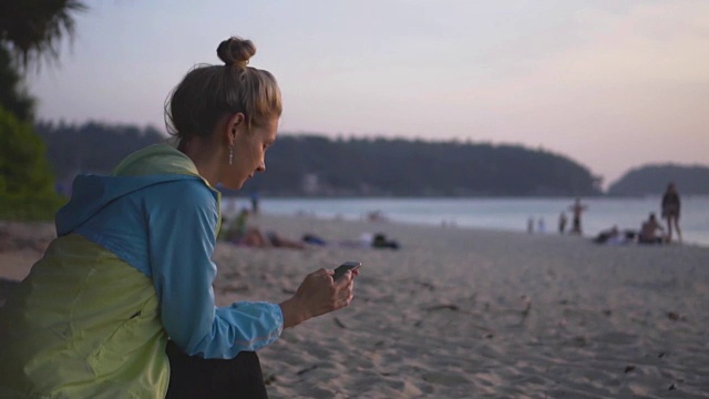 日落时分，一个拿着手机的悲伤女孩坐在海滩上。视频下载