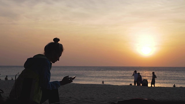 夕阳西下时，一个悲伤的女孩坐在海滩上写着电话短信。视频下载