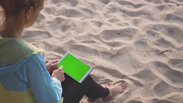 一个女孩坐在沙滩上，用着带有彩色按键屏幕的平板电脑。刷。视频下载