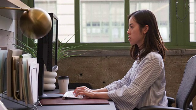 一位年轻的亚洲女性在办公室里使用电脑，侧面视频素材
