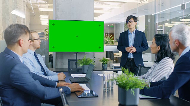 亚洲商人向他的商业伙伴做报告/报告，指着绿色度键屏幕墙电视。视频素材