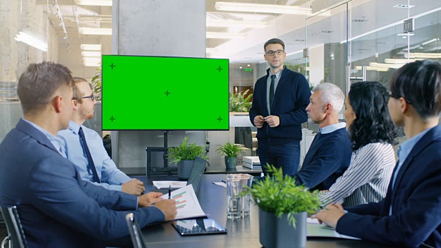 年轻的天才商人给他的商业同事报告/报告，指着绿色色度键屏幕墙电视。视频素材