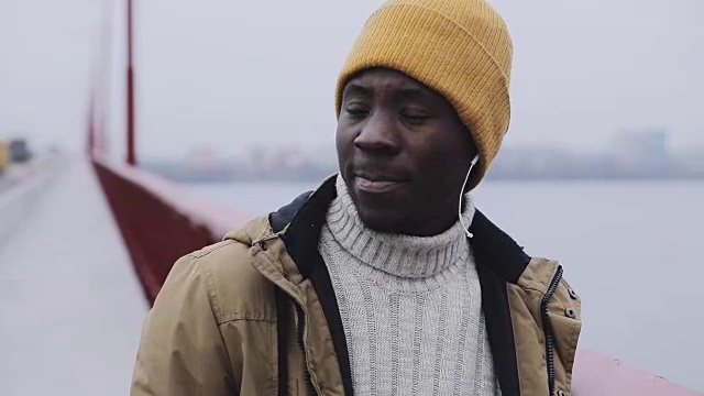 年轻的黑色微笑男子肖像在寒冷的冬天站在一座桥上听耳机视频素材