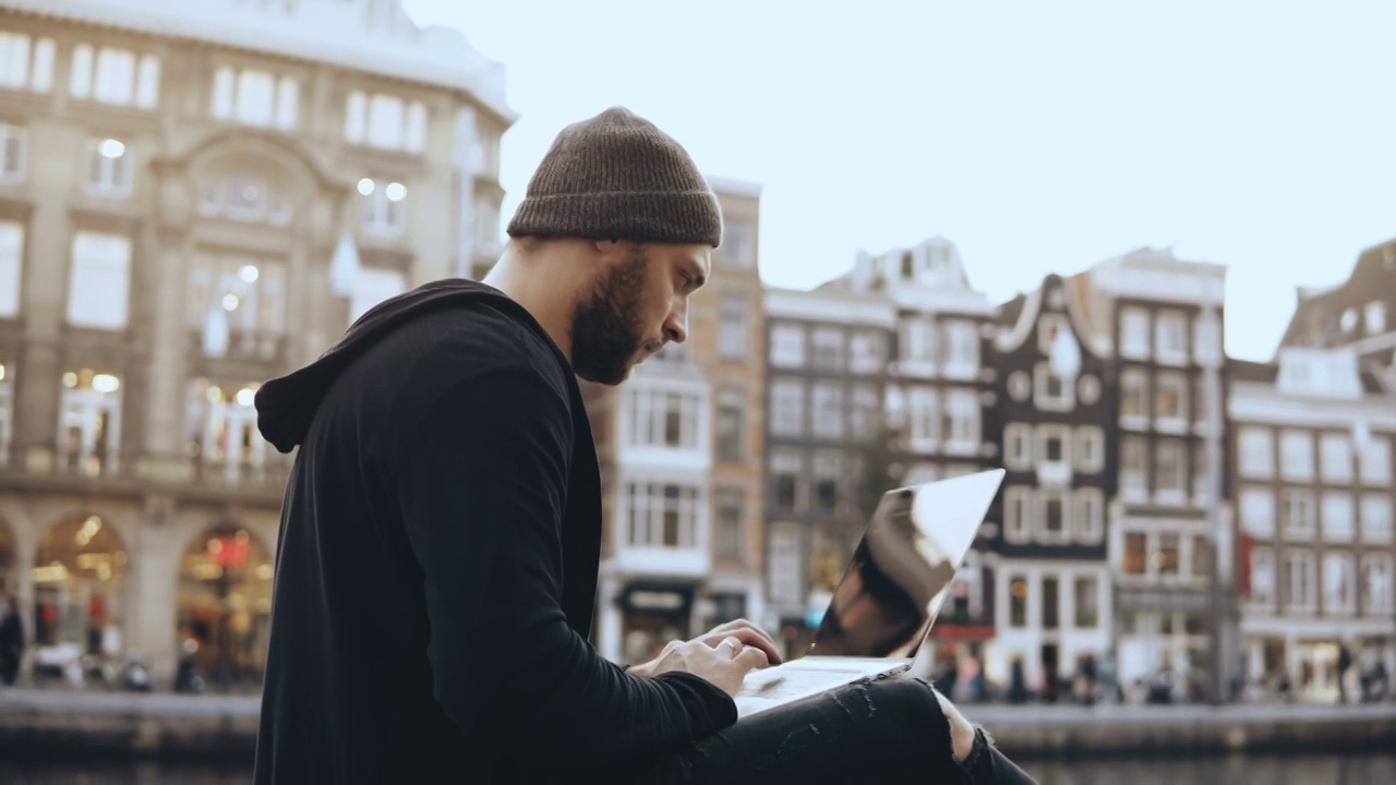 一个有创造力的人坐在街上，拿着笔记本电脑。移动办公室工作。阿姆斯特丹老城。大气的城市视频下载