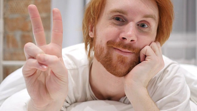 年轻的红发胡子男子的胜利手势，在床上的积极姿态视频素材