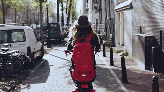 后视图。女性在阳光城市骑自行车。缓慢的运动。女孩在时尚的帽子和红色背包在自行车上。探索视频素材