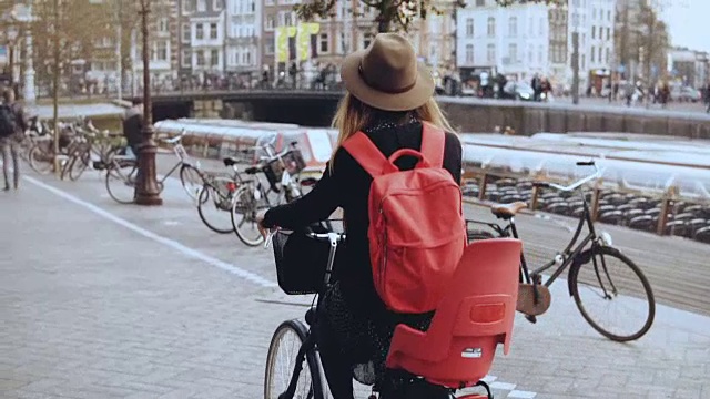 女人在拥挤的老城区街道上骑自行车。后视图慢动作。女性骑在自行车上戴着帽子和红色背包视频素材
